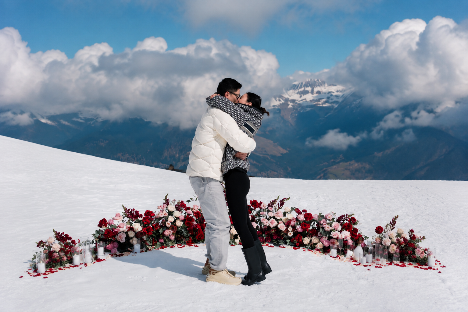 Couple kissing in a snowy mountain proposal surrounded by a heart-shaped arrangement of red and pink flowers